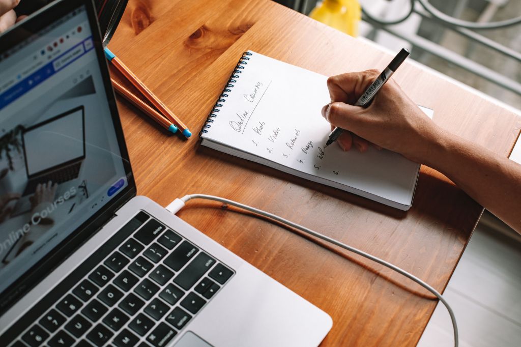 A laptop open at a online course homepage. Next to the laptop is a hand taking notes using paper and pen. Image by Roman Odintsov.